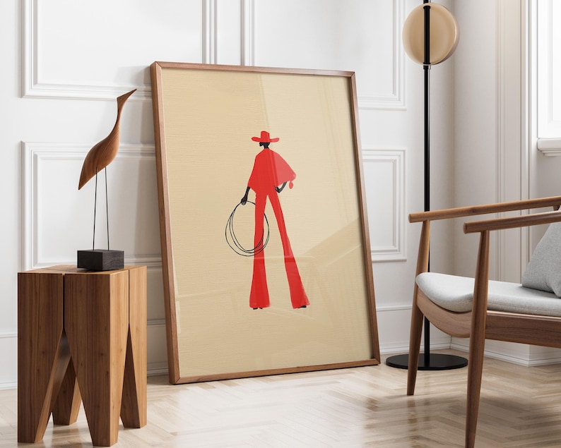 May include: A framed art print featuring a stylized illustration of a person in a red outfit holding a lasso. The artwork has a beige background and a wooden frame. A wooden sculpture of a bird and a wooden side table are in the foreground.