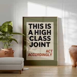 May include: A framed poster with the text "THIS IS A HIGH CLASS JOINT ACT ACCORDINGLY" in black and red. The poster is in a room with a plant in a terracotta pot, a gray ottoman, and decorative vases.