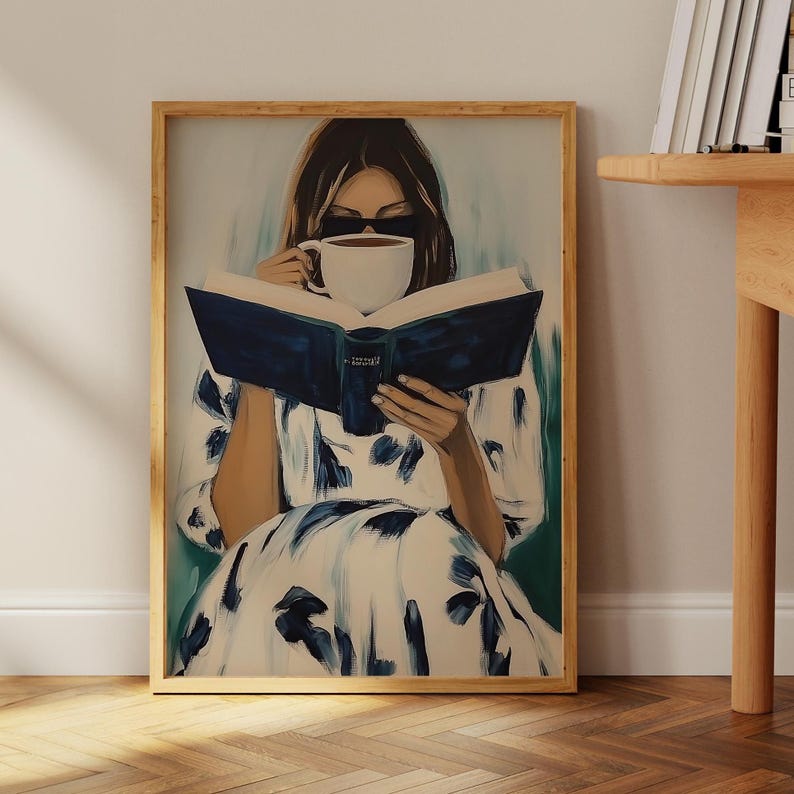 May include: A framed art print depicts a person reading a book and drinking from a white mug. The person is wearing sunglasses and a white dress with blue floral patterns. The book has a dark blue cover.