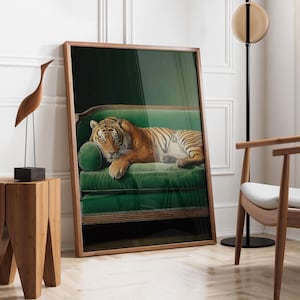 May include: A framed art print depicting a tiger reclining on a green velvet sofa. The tiger has orange and black stripes. The sofa has a dark wooden frame. The artwork is leaning against a white wall. A wooden sculpture and chair are in the background.