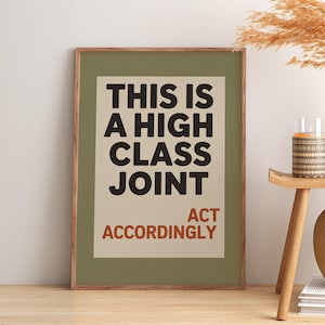 May include: A framed art print with the text "THIS IS A HIGH CLASS JOINT ACT ACCORDINGLY" in black and red. The print is in a green frame and is displayed on a wooden surface. A small table with a candle and vase is next to the print.