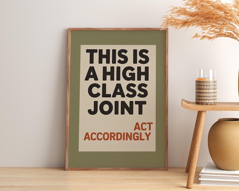 May include: A framed art print with the text "THIS IS A HIGH CLASS JOINT ACT ACCORDINGLY" in black and red. The print is in a green frame and is displayed on a wooden surface. A small table with a candle and vase is next to the print.