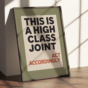 May include: Framed poster with the text "THIS IS A HIGH CLASS JOINT ACT ACCORDINGLY" in black and red on an off-white background. The frame is dark green, and the poster is leaning against a white wall on a wooden floor.