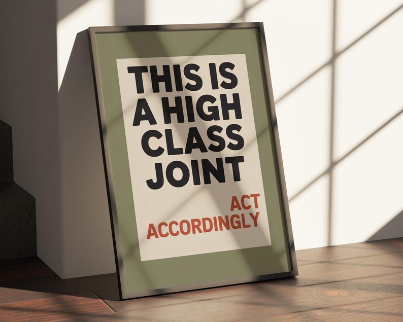 May include: Framed poster with the text "THIS IS A HIGH CLASS JOINT ACT ACCORDINGLY" in black and red on an off-white background. The frame is dark green, and the poster is leaning against a white wall on a wooden floor.