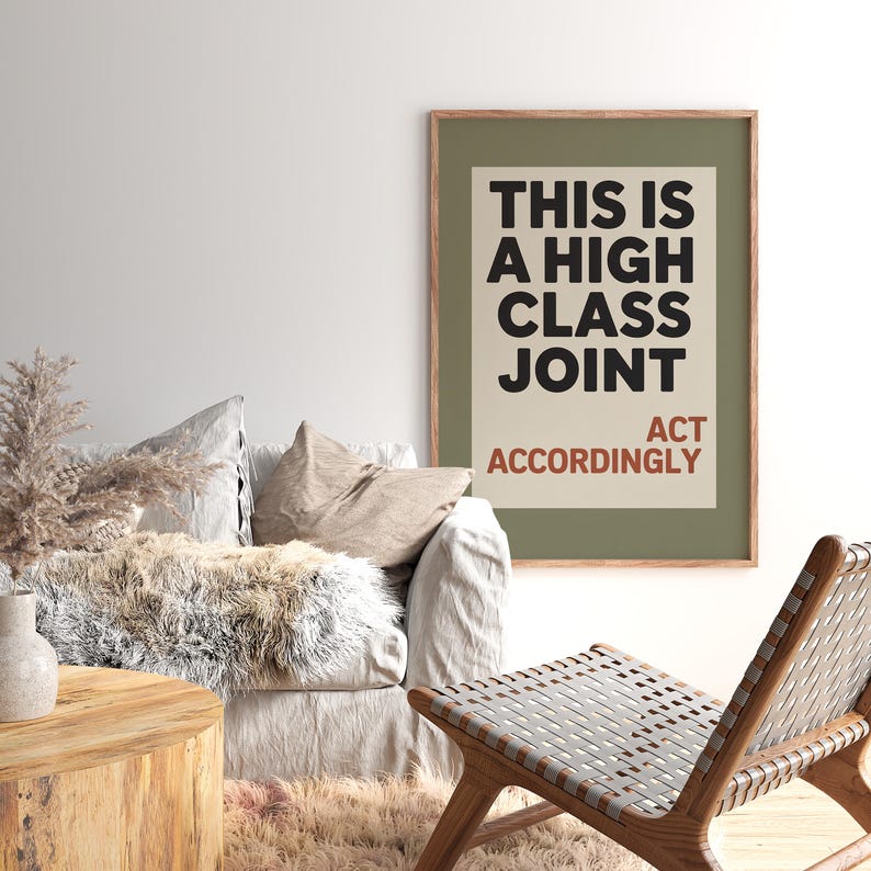 May include: A framed art print with the text "THIS IS A HIGH CLASS JOINT ACT ACCORDINGLY" in black and red. The print is displayed above a sofa with pillows and a faux fur throw. A wooden chair is in the foreground.