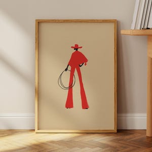 May include: A framed art print featuring a stylized illustration of a person in a red cowboy outfit, holding a lasso. The figure is set against a beige background, with a wooden frame. The artwork has a minimalist aesthetic.