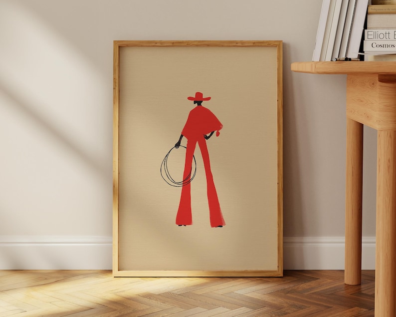 May include: A framed art print featuring a stylized illustration of a person in a red cowboy outfit, holding a lasso. The figure is set against a beige background, with a wooden frame. The artwork has a minimalist aesthetic.