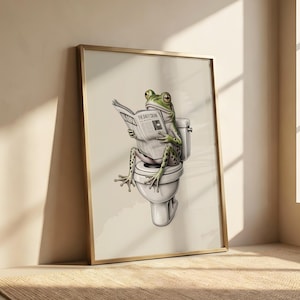 May include: A framed art print depicting a green frog reading a newspaper titled "The Daily Croak" whilst seated on a white toilet. The frog has black spots and is in a relaxed pose. The frame is gold.