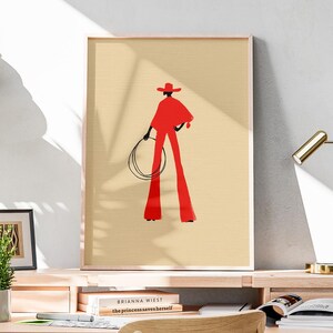 May include: A framed art print featuring a stylized illustration of a person in a red outfit, holding a lasso. The figure is set against a beige background. The artwork is displayed on a wooden desk with a desk lamp and books.