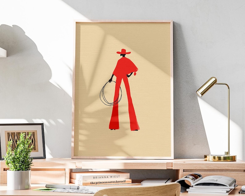 May include: A framed art print featuring a stylized illustration of a person in a red outfit, holding a lasso. The figure is set against a beige background. The artwork is displayed on a wooden desk with a desk lamp and books.