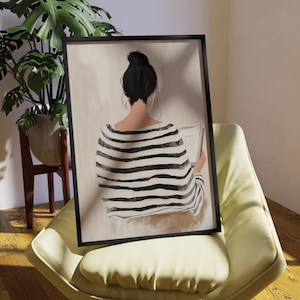 May include: A framed illustration of a person sitting and reading a book. The person is wearing a black and white striped shirt and has dark hair. The background is a light beige color.