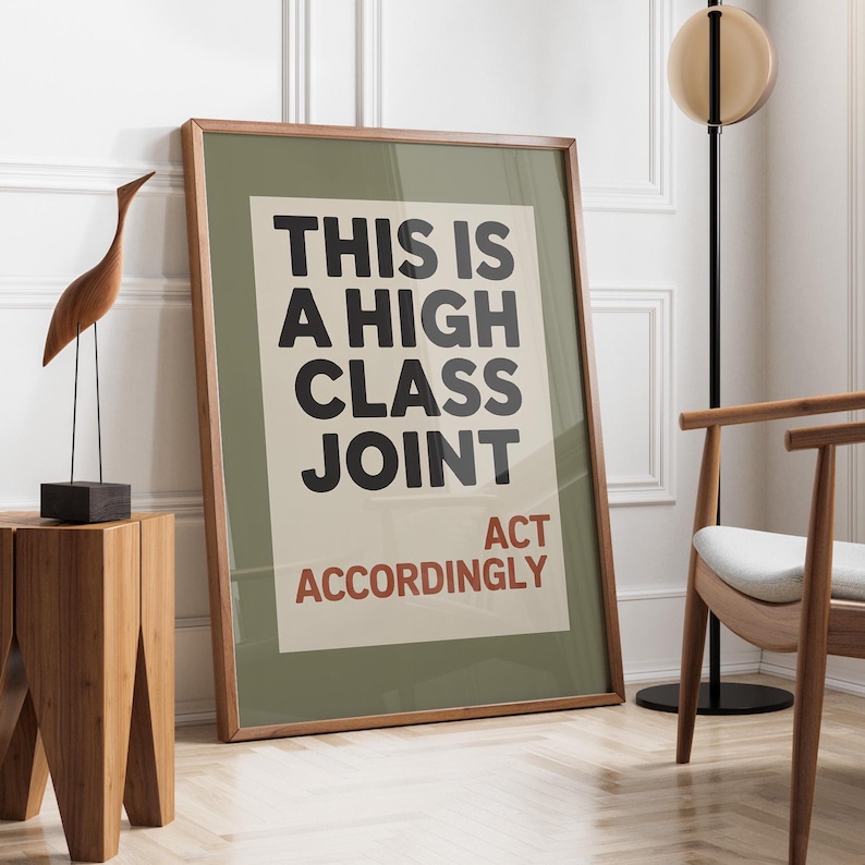 May include: A framed poster with the text "THIS IS A HIGH CLASS JOINT ACT ACCORDINGLY" in black and red. The poster is in a room with a wooden side table, a wooden bird sculpture, and a modern chair.