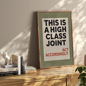May include: A framed art print with the text "THIS IS A HIGH CLASS JOINT ACT ACCORDINGLY" in black and red. The print is in a green frame and rests on a wooden cabinet. A white cat figurine and books are on the cabinet.