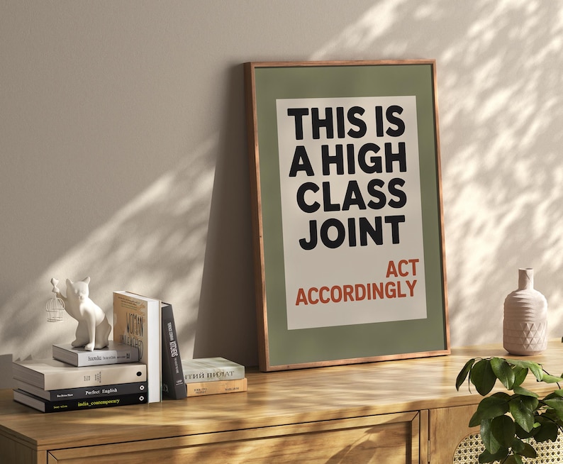May include: A framed art print with the text "THIS IS A HIGH CLASS JOINT ACT ACCORDINGLY" in black and red. The print is in a green frame and rests on a wooden cabinet. A white cat figurine and books are on the cabinet.