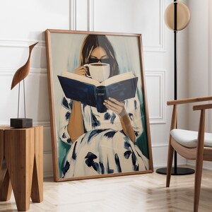 May include: A framed art print depicts a person reading a book and drinking from a white mug. The artwork features a woman in a white dress with blue floral patterns. A wooden sculpture of a bird and a small wooden table are in the foreground.