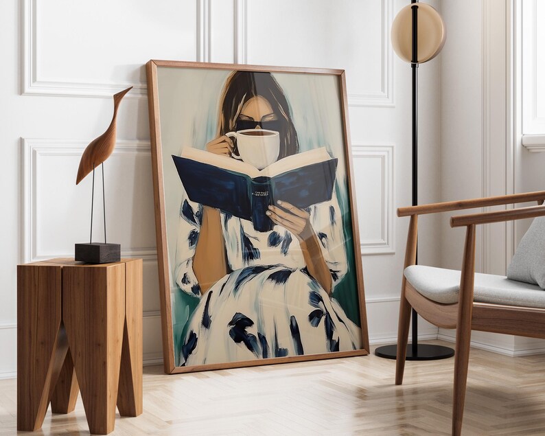 May include: A framed art print depicts a person reading a book and drinking from a white mug. The artwork features a woman in a white dress with blue floral patterns. A wooden sculpture of a bird and a small wooden table are in the foreground.