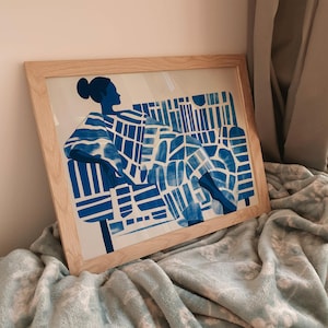 Op de afbeelding: Een ingelijste illustratie van een persoon die op een stoel zit. De persoon en de stoel zijn beide blauw en wit met een geometrisch patroon.