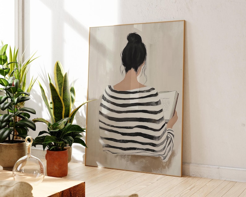 May include: A black and white striped shirt with a woman sitting and reading a book. The woman has her back to the viewer and her hair is pulled back in a bun. The background is a light beige color.