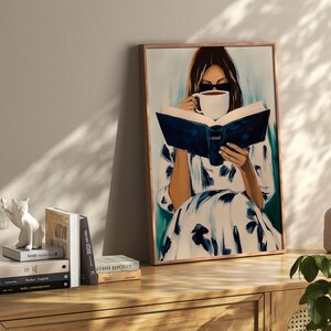 May include: A framed painting depicts a person reading a book and drinking coffee. The person is wearing a white dress with blue floral patterns. The painting is displayed on a wooden cabinet, with books and decorative items.