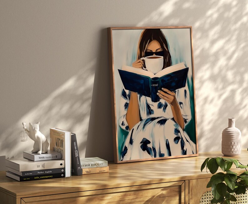 May include: A framed painting depicts a person reading a book and drinking coffee. The person is wearing a white dress with blue floral patterns. The painting is displayed on a wooden cabinet, with books and decorative items.