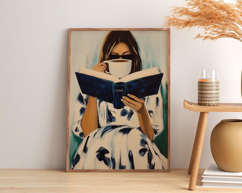 May include: A framed art print depicts a person reading a book while holding a cup of coffee. The person is wearing sunglasses and a white dress with blue floral patterns. The artwork has a soft, painterly style with a neutral color palette.