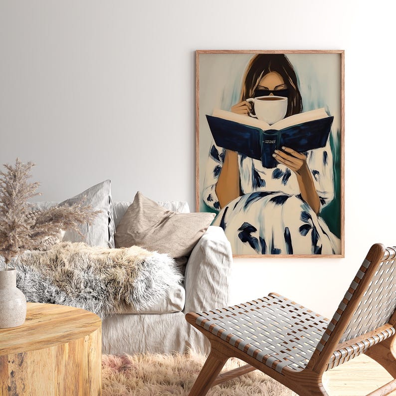 May include: A framed art print depicts a person reading a book and drinking from a white mug. The person is wearing a white dress with blue floral patterns. The book has a dark blue cover. The art has a light brown frame.
