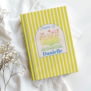 Pode incluir: Um caderno personalizado com uma capa listrada amarela e branca. A capa apresenta um design floral acima do nome "Danielle" em azul. O texto "Capítulo 18" está arqueado acima do design floral. O caderno está em uma superfície branca.