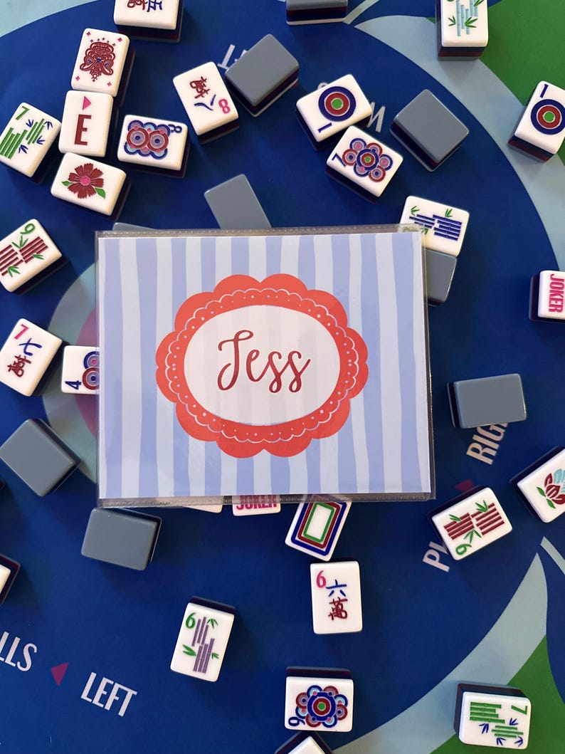 May include: A collection of Mahjong tiles and gray tile holders scattered on a blue surface. A card with the name "Jess" in a red and white design is in the center. The tiles have various symbols and numbers.