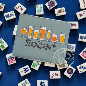May include: A light blue Mahjong tile holder with the name "Robert" and a beer graphic. The holder is surrounded by white Mahjong tiles on a blue surface. The tiles have various symbols and numbers.