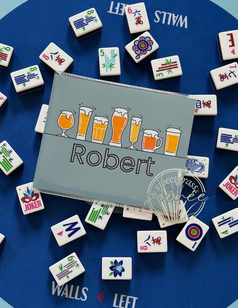 May include: A light blue Mahjong tile holder with the name "Robert" and a beer graphic. The holder is surrounded by white Mahjong tiles on a blue surface. The tiles have various symbols and numbers.