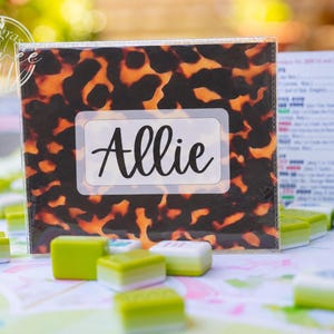 Può includere: Un porta carte rettangolare in plastica con motivo tartarugato marrone e nero, dotato di un'etichetta centrale bianca che riporta il nome Allie in corsivo nero, posizionato tra diverse piccole tessere da gioco verdi e bianche.