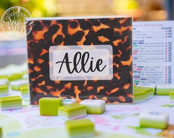 May include: A rectangular plastic card holder featuring a brown and black tortoiseshell pattern with a white center label displaying the name Allie in black script. The holder is positioned among several small green and white game tiles.