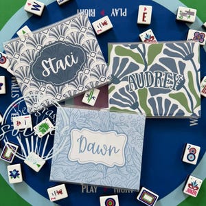 May include: Three personalized Mahjong tile cases with names "Staci", "Audrey", and "Dawn" on them. The cases have different floral and geometric patterns in blue, green, and white. Mahjong tiles surround the cases on a blue and green surface.