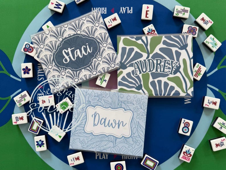 May include: Three personalized Mahjong tile cases with names "Staci", "Audrey", and "Dawn" on them. The cases have different floral and geometric patterns in blue, green, and white. Mahjong tiles surround the cases on a blue and green surface.
