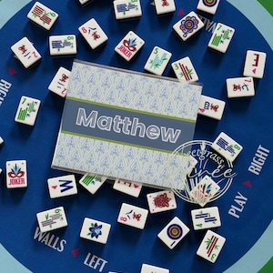 May include: A blue Mahjong game mat with scattered tiles. A personalized card with the name "Matthew" is in the center. The tiles are white with various symbols and numbers. The mat has text that says "Walls", "Left", "Play", and "Right".