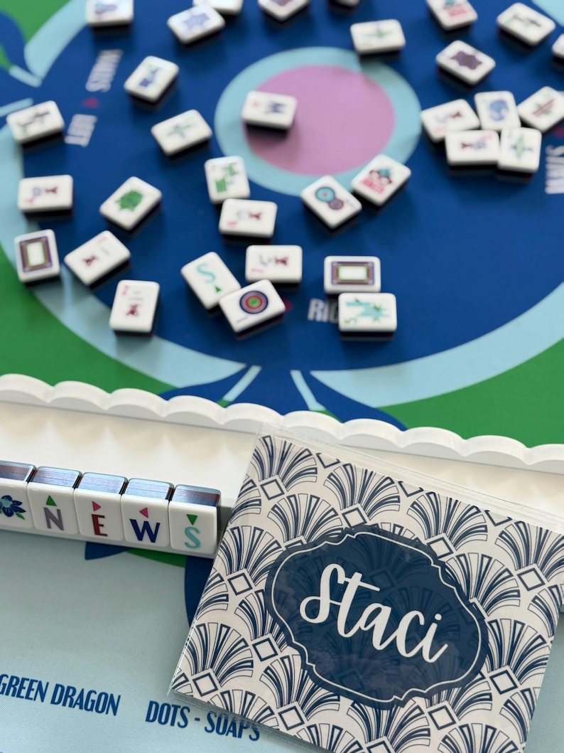 May include: A tabletop game with white tiles scattered on a blue game board. The tiles have various symbols and designs. A small bag with the name "Staci" is on the right. The word "NEWS" is spelled out with tiles.