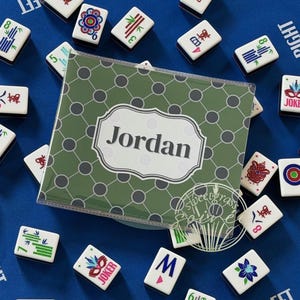 May include: A blue game board with scattered Mahjong tiles and a personalized green case that reads "Jordan". The tiles are white with various symbols and numbers. The case has a green and gray pattern.