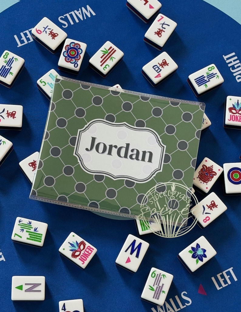 May include: A blue game board with scattered Mahjong tiles and a personalized green case that reads "Jordan". The tiles are white with various symbols and numbers. The case has a green and gray pattern.