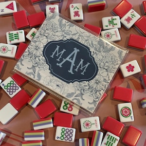 May include: A collection of red and white Mahjong tiles with various symbols and numbers scattered on a pink surface. A card with the initials "MAM" in a floral design is also present. The tiles are a mix of red, white, and multi-colored.