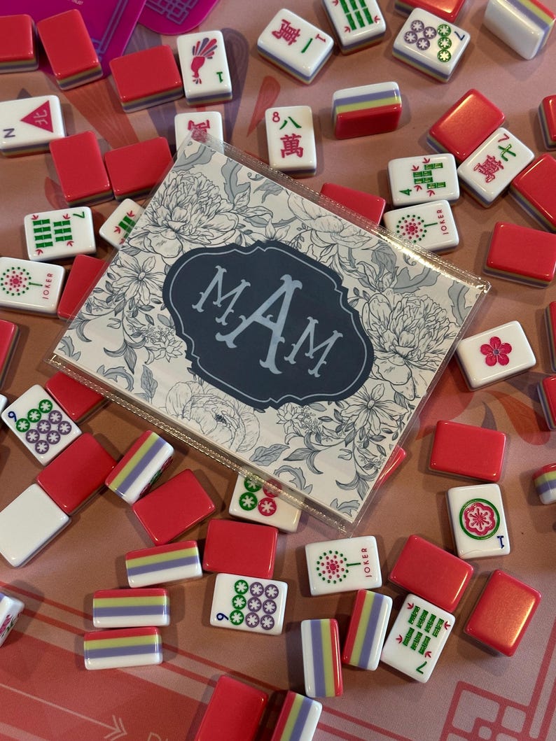 May include: A collection of red and white Mahjong tiles with various symbols and numbers scattered on a pink surface. A card with the initials "MAM" in a floral design is also present. The tiles are a mix of red, white, and multi-colored.
