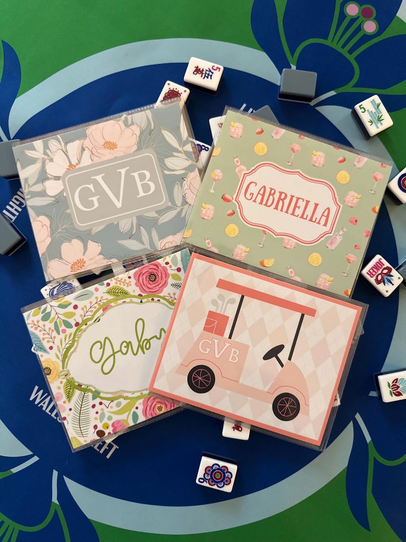 May include: Four personalized coasters with floral and golf-themed designs. One coaster features the initials "GVB" in a gray box, another has the name "Gabriella" in a decorative frame, and a third has the name "Gabi". A fourth coaster has a pink golf cart.