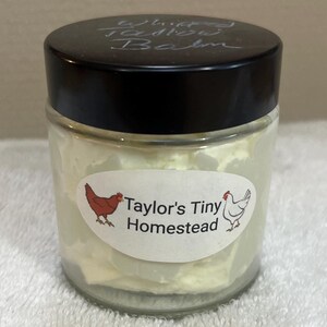 Puede incluir: Un tarro de vidrio con tapa negra lleno de bálsamo corporal batido blanco. El tarro tiene una etiqueta con el texto "Taylor's Tiny Homestead" e imágenes de una gallina roja y una gallina blanca.