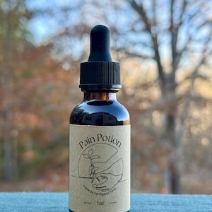 Puede incluir: Una botella de vidrio marrón con un cuentagotas negro. La etiqueta dice "Pain Potion" con un gráfico de una mano sosteniendo una planta. La etiqueta también dice "made by Earthganic Farm" y "1 oz".