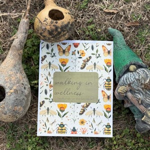 May include: A journal with the title "walking in wellness" and a cover design of butterflies, bees, and flowers. The image also includes two gourd birdhouses and a garden gnome. The journal rests on grass and dry leaves.
