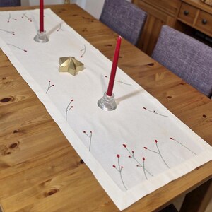 May include: A white table runner with a delicate embroidered design of red berries and branches. Two red candlesticks are placed on the runner, adding a touch of elegance to the table setting.