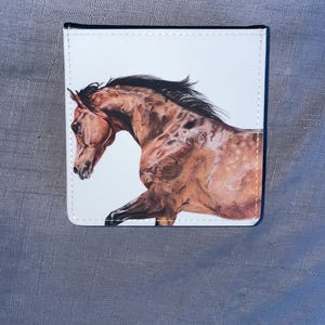 May include: A white wallet with a detailed illustration of a brown horse in full gallop. The horse has a black mane and is set against a white background. The wallet has a black border and is likely made of leather or a similar material.