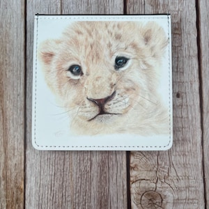 May include: White wallet with a detailed illustration of a lion cub's face. The cub has light brown fur, blue eyes, and a small nose. The wallet has a black border and is set against a wooden background.