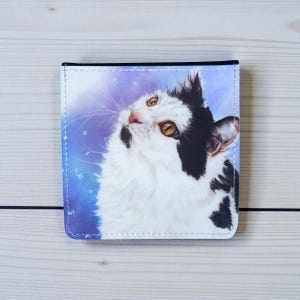 May include: A wallet featuring a white and black cat illustration against a blue and purple backdrop. The wallet has a white border, black trim, and a stitched edge, depicting a cat looking upwards.