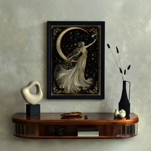 May include: A framed artwork shows a woman in a flowing gown reaching for a star, set against a crescent moon and a starry night. The artwork is framed in black and gold. The image is displayed on a wooden shelf.