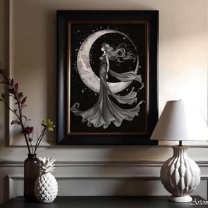 May include: A framed black and white print depicts a woman standing on a crescent moon against a starry background. The artwork is in a black frame with a gold inner border. A white lamp with a textured base sits on a dark surface.